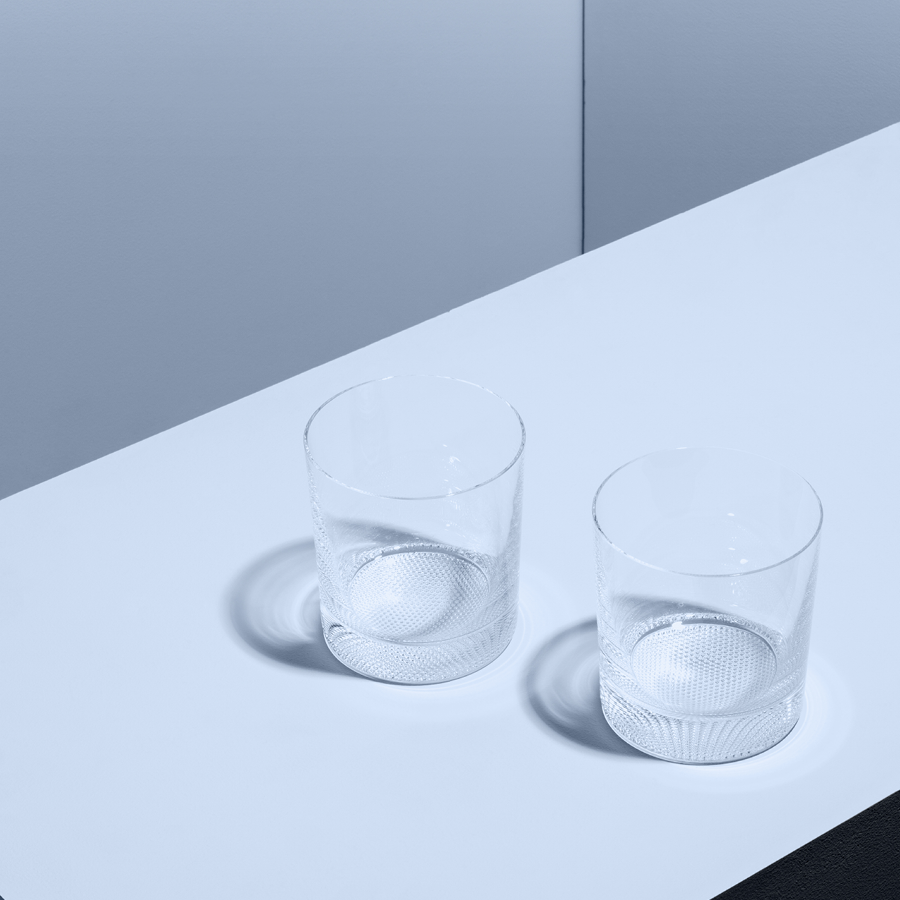 Two KostaBoda Limelight Double Old Fashioned Glasses, each 15oz, rest elegantly on a white tabletop, casting soft shadows against the light gray background. The clear glass tumblers minimalist design exudes sophistication.