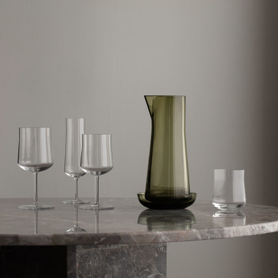 An Orrefors glass carafe and four stylish 9.5oz Informal Glasses (2-pack), echoing Scandinavian design akin to Björn Dahlströms style, are elegantly arranged on a round marble table against a plain gray wall.