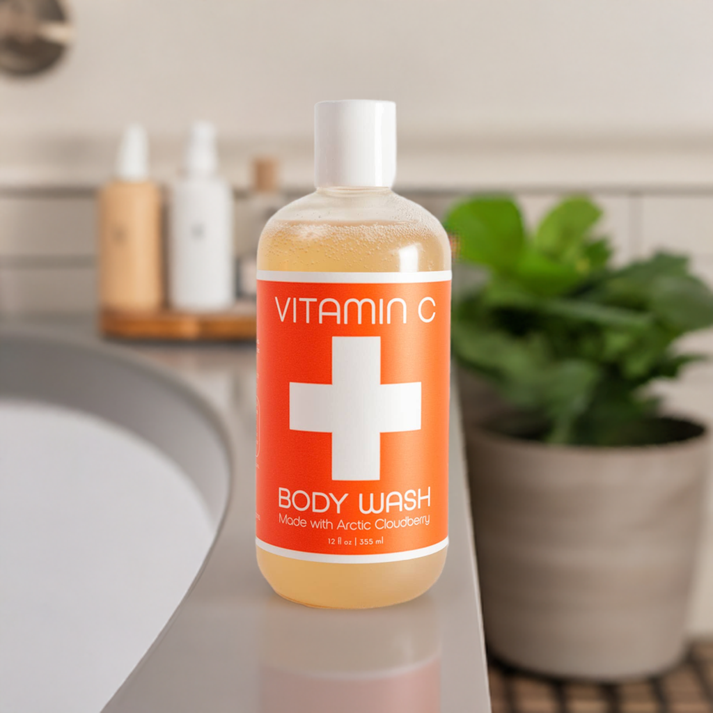 A bottle of Nordic+Wellness™ Vitamin C Body Wash (12 fl oz) with an orange label and enhanced by Arctic Cloudberry Oil, featuring a white cross, sits on a bathroom counter. A plant and toiletries are blurred in the background.