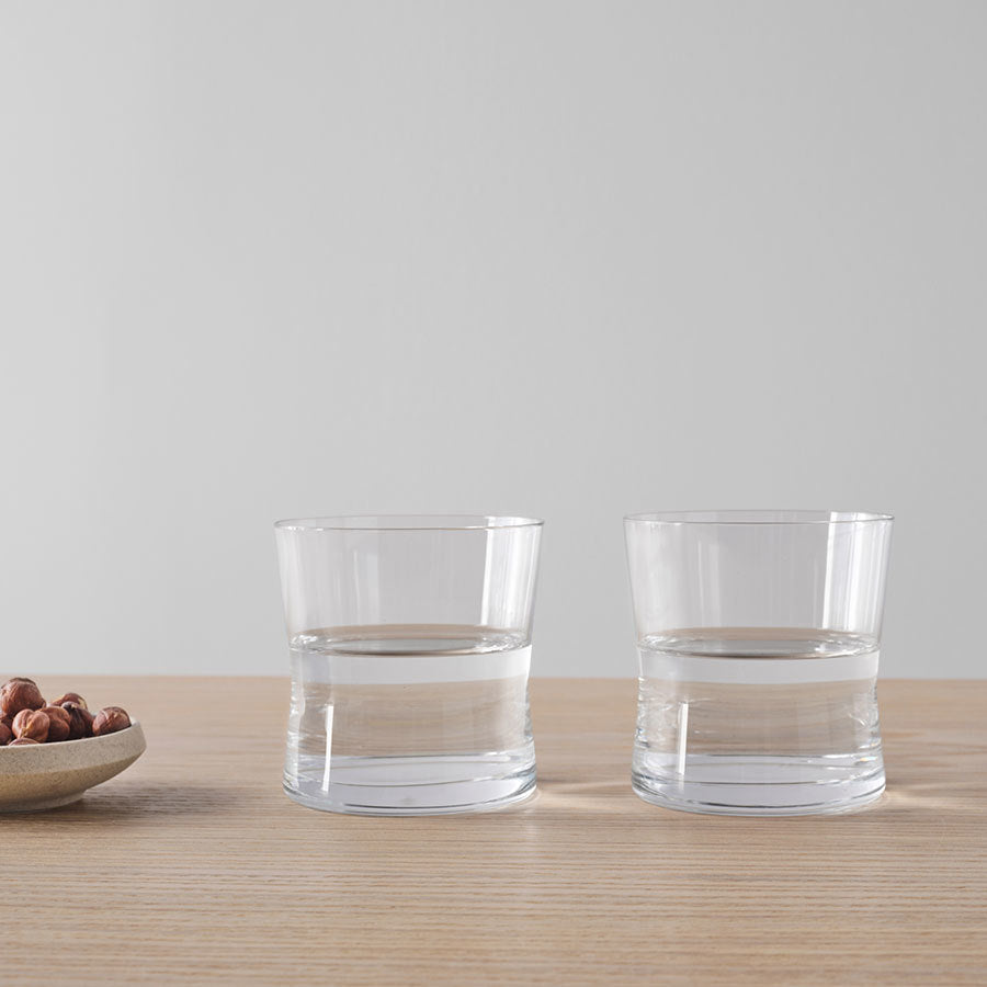 Two Orrefors Grace Double Old Fashioned 13.2oz glasses of water are on a wooden table beside a small plate of hazelnuts, set against a plain gray wall that highlights Erika Lagerbielkes elegant design.