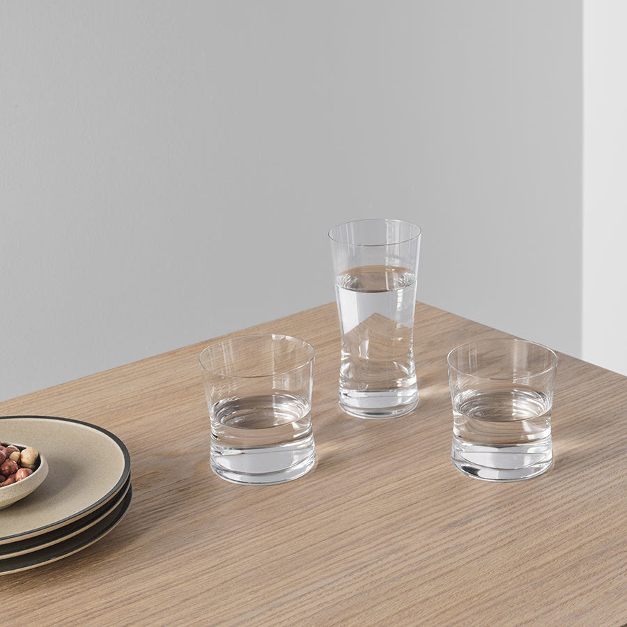 On the wooden table, two empty glasses sit alongside a filled glass of water, stacked plates, and a dish with nuts. Among them is an elegant Orrefors Grace Double Old Fashioned 13.2oz from the renowned designer Erika Lagerbielke.