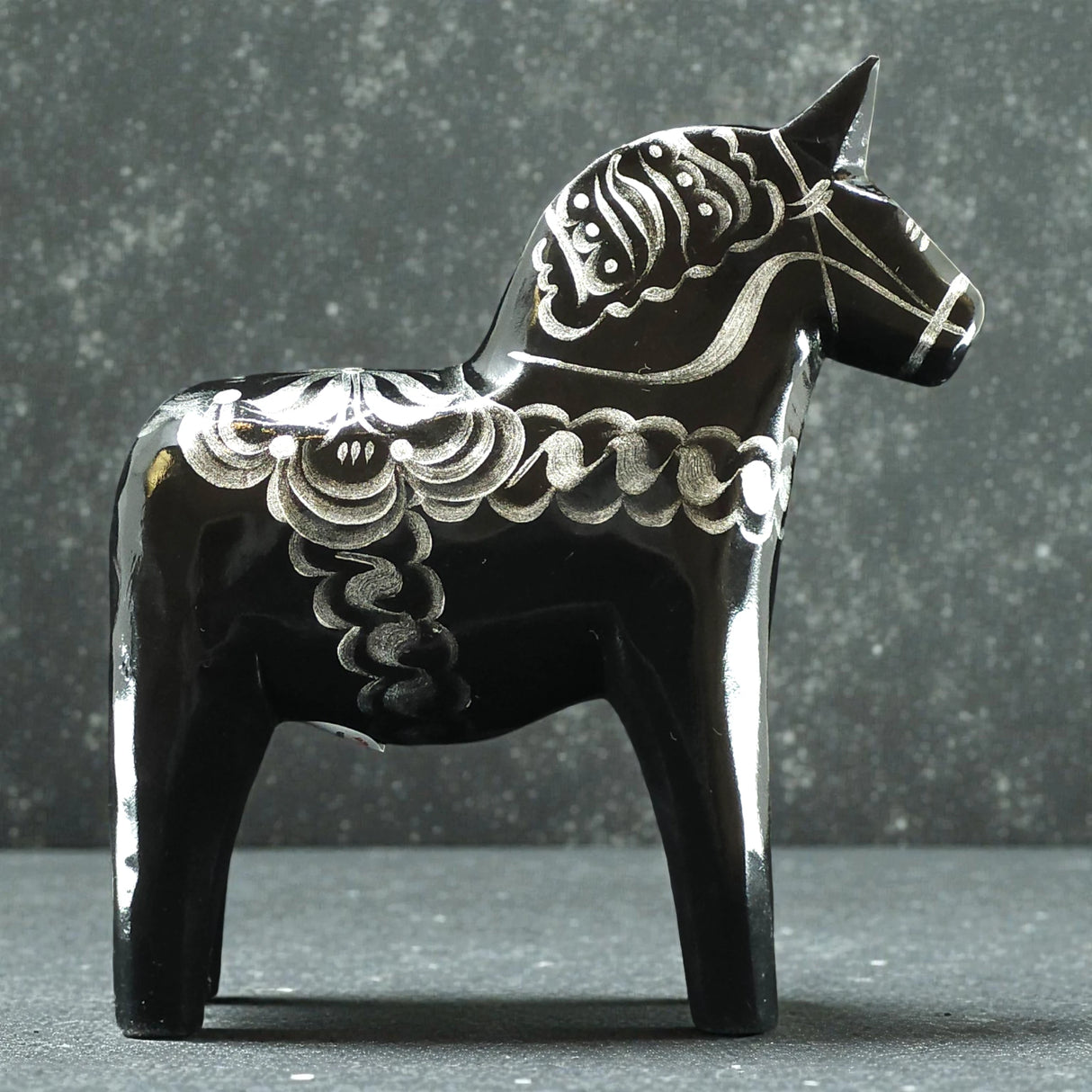 The Black & Silver Swedish Dala Horse "Celebrate Series" by Grannas Olsson, handcrafted in Sweden (13 cm), stands on a speckled surface against a gray backdrop, showcasing the charm of Swedish folk art.