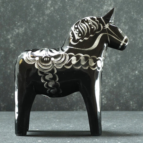 The Black & Silver Swedish Dala Horse "Celebrate Series" by Grannas Olsson, handcrafted in Sweden (13 cm), stands on a speckled surface against a gray backdrop, showcasing the charm of Swedish folk art.