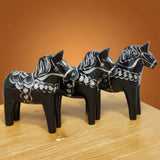 Three black & silver "Celebrate Series" Dala Horses by Grannas Olsson, each handcrafted in Sweden (13 cm / 5 in), stand together on a light wooden surface against a warm orange-brown background.