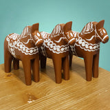 Three handcrafted 13 cm brown "Gingerbread" Dala Horses by Grannas Olsson, featuring white Scandinavian patterns, stand on a wooden surface against a teal background.