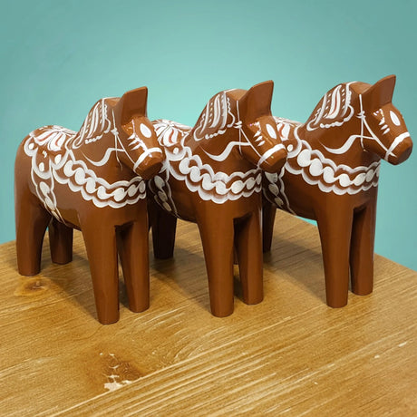Three handcrafted 13 cm brown "Gingerbread" Dala Horses by Grannas Olsson, featuring white Scandinavian patterns, stand on a wooden surface against a teal background.