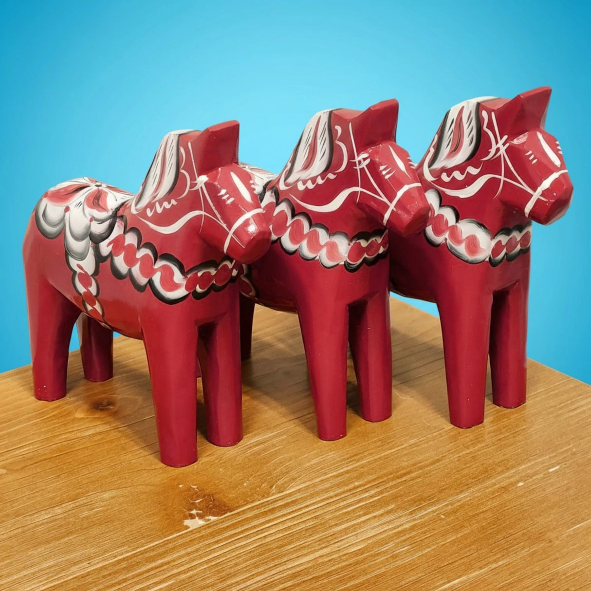 Three handcrafted Grannas Olsson "Maria / Anniversary Series" Burgundy Swedish Dala Horses (15 cm), featuring white and black details, stand on a wooden surface against a blue background.
