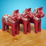 Three handcrafted Grannas Olsson "Maria / Anniversary Series" Burgundy Swedish Dala Horses (15 cm), featuring white and black details, stand on a wooden surface against a blue background.