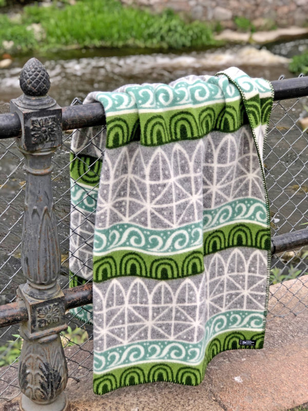 The Wool Blanket: Uppsala, Grey, Green, White is draped over a metal railing beside a flowing river in Uppsala, surrounded by grass and stones.