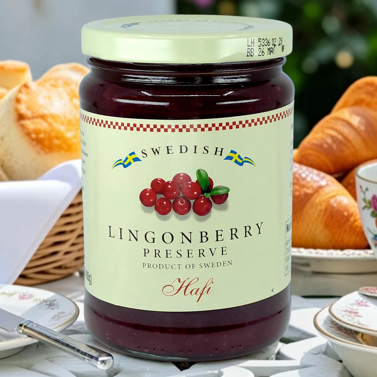 A jar of Swedish lingonberry preserves on a table with bread rolls, a croissant, a knife, and a floral teacup in the background.