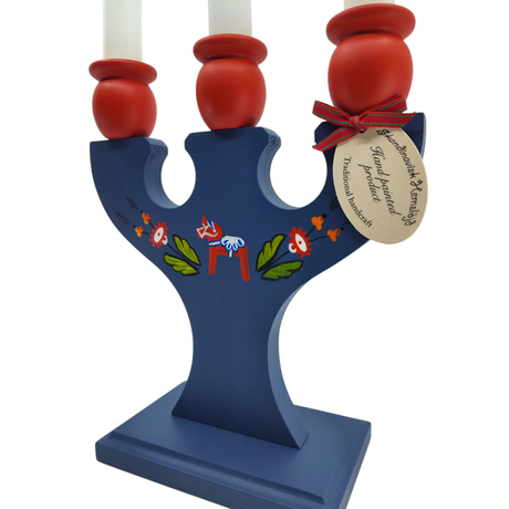The Candle Holder is a handcrafted Swedish piece featuring a Dala Horse and kurbits, designed with blue wood and floral motifs. It holds three candles and includes a tag attached by a red ribbon.