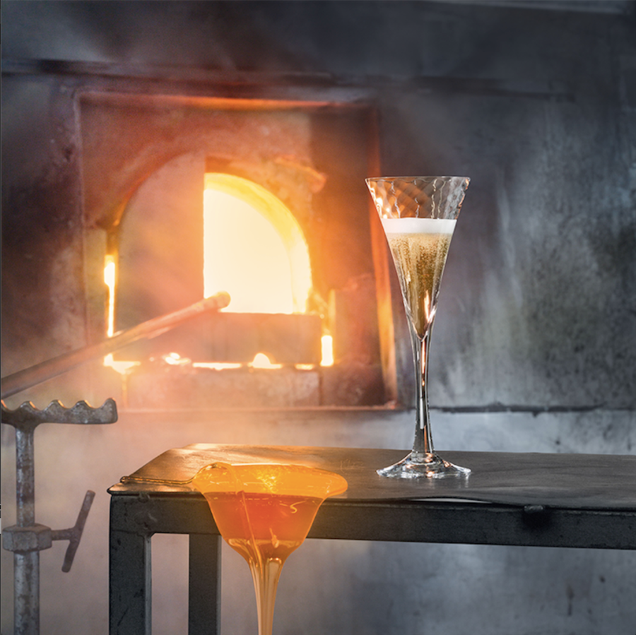 An Orrefors: Helena Champagne Glass 8.5oz from the (4-Pack) sits on a metal table by a glassblowing furnace, its warm glow reflecting the craftsmanship reminiscent of Gunnar Cyréns designs.