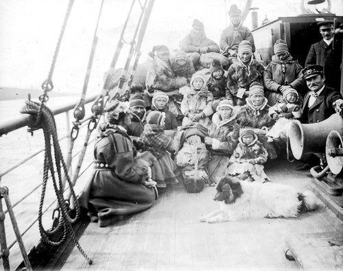 A historical black-and-white photo captures a group in warm clothing on a ship's deck, echoing the poignant story from "Book: Rocks will Echo Our Sorrow The Forced Displacement of the Northern Sami." The presence of Northern Sámi community members and a dog highlights Elin Anna Labba's narratives of resilience and cultural heritage.