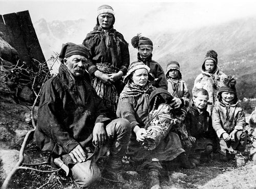 A historical black and white photo captures a group of Northern Sámi people, including adults and children, wearing traditional clothing. They stand outdoors in a mountainous area, evoking poignant memories of Sámi displacement eloquently chronicled in Elin Anna Labba's book *Rocks will Echo Our Sorrow: The Forced Displacement of the Northern Sami*.