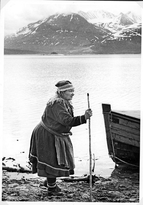 Dressed in traditional Northern Sámi attire, a person stands by a boat on a rocky shore, stick in hand. Snow-capped mountains and a tranquil lake form the backdrop, mirroring the poignant narratives of Sámi displacement explored in the book "Rocks will Echo Our Sorrow: The Forced Displacement of the Northern Sami" by Elin Anna Labba.