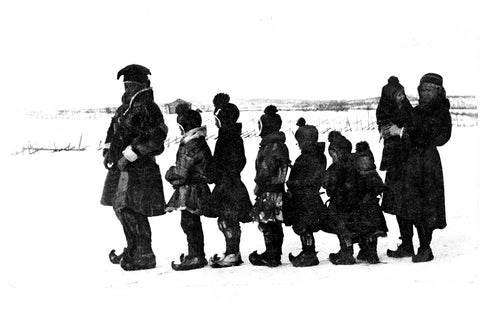 A line of people, including children dressed in winter attire, walk single file through the snowy landscape, evoking scenes from "Rocks will Echo Our Sorrow: The Forced Displacement of the Northern Sámi." A woman at the end carries a baby, reflecting the hardships faced during Sámi displacement.