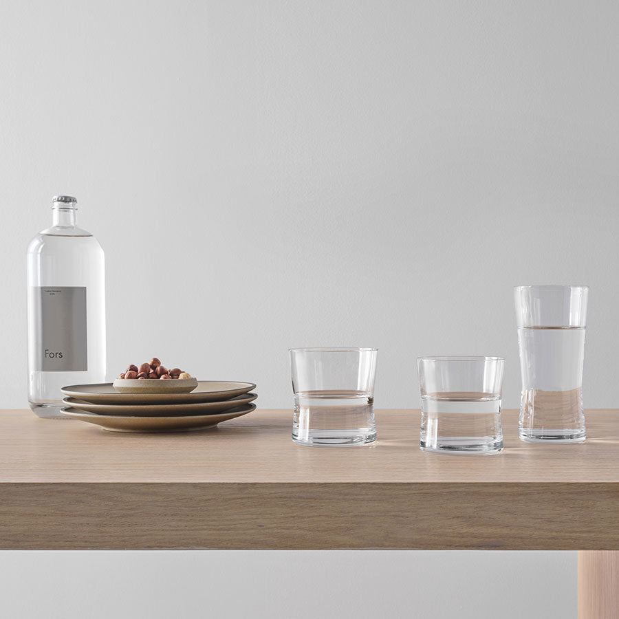 A wooden table displays three stacked plates, a bowl of nuts, and a water bottle next to three Orrefors Grace Highball Glasses, 14.5oz (2-Pack), filled with water and elegantly designed by Erika Lagerbielke.