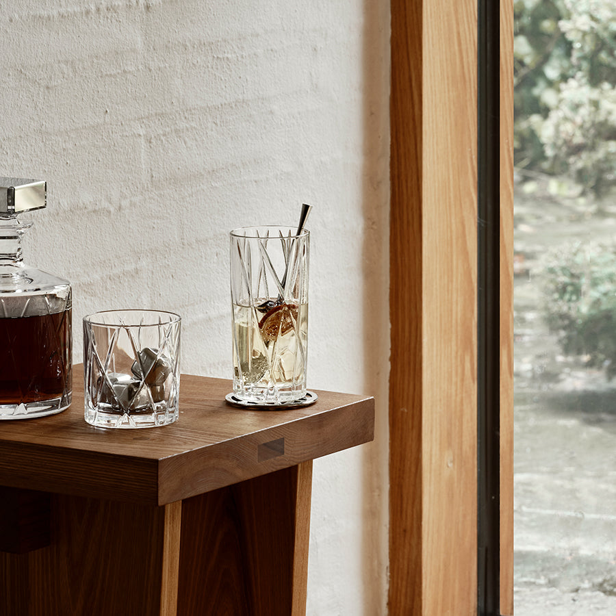 Two glasses with drinks sit on a wooden table by a window: one is a City Highball Glass from Orrefors, decorated with stones next to a decanter; the other is also an Orrefors City Highball Glass, filled with ice and topped with a straw.