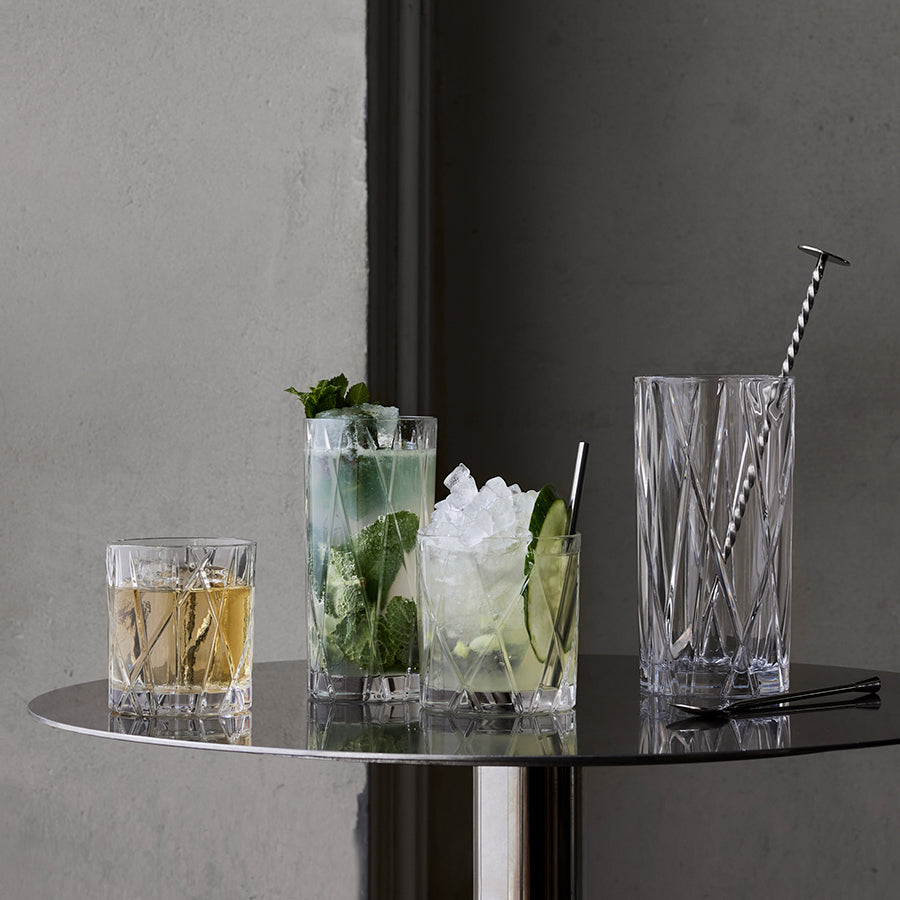 Two packs of Orrefors City Highball 12oz glasses with varied drinks and a sleek cocktail mixer adorn a round table against a gray backdrop.