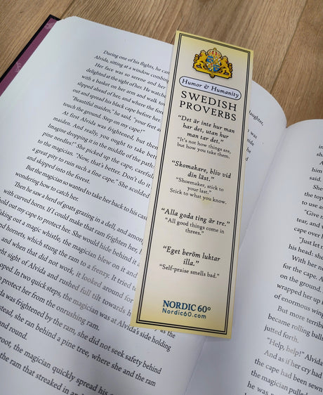 The “Humor & Humanity” Swedish Proverbs Bookmark (2.5" × 8.5") rests on an open book, featuring five proverbs in both Swedish and English, inspired by Scandinavian tradition and perfect for any wooden surface.