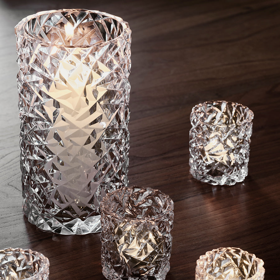 The Orrefors Carat Hurricane candle holders, with one large and several small faceted glass pieces resembling a cut crystal vase, sit on a wooden surface, lit from within.