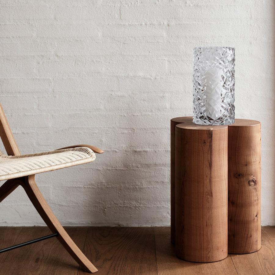 A wooden chair and a stump side table with an Orrefors Carat Hurricane vase sit against a white brick wall.