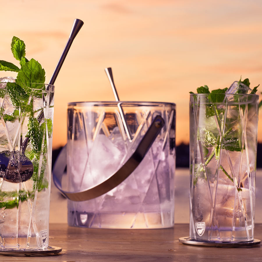 Three crystal glasses with mint leaves, ice, and straws adorn a table, centered around the transparent splendor of the Orrefors: City Ice Bucket. The scene is complemented by a breathtaking sunset sky in the background.