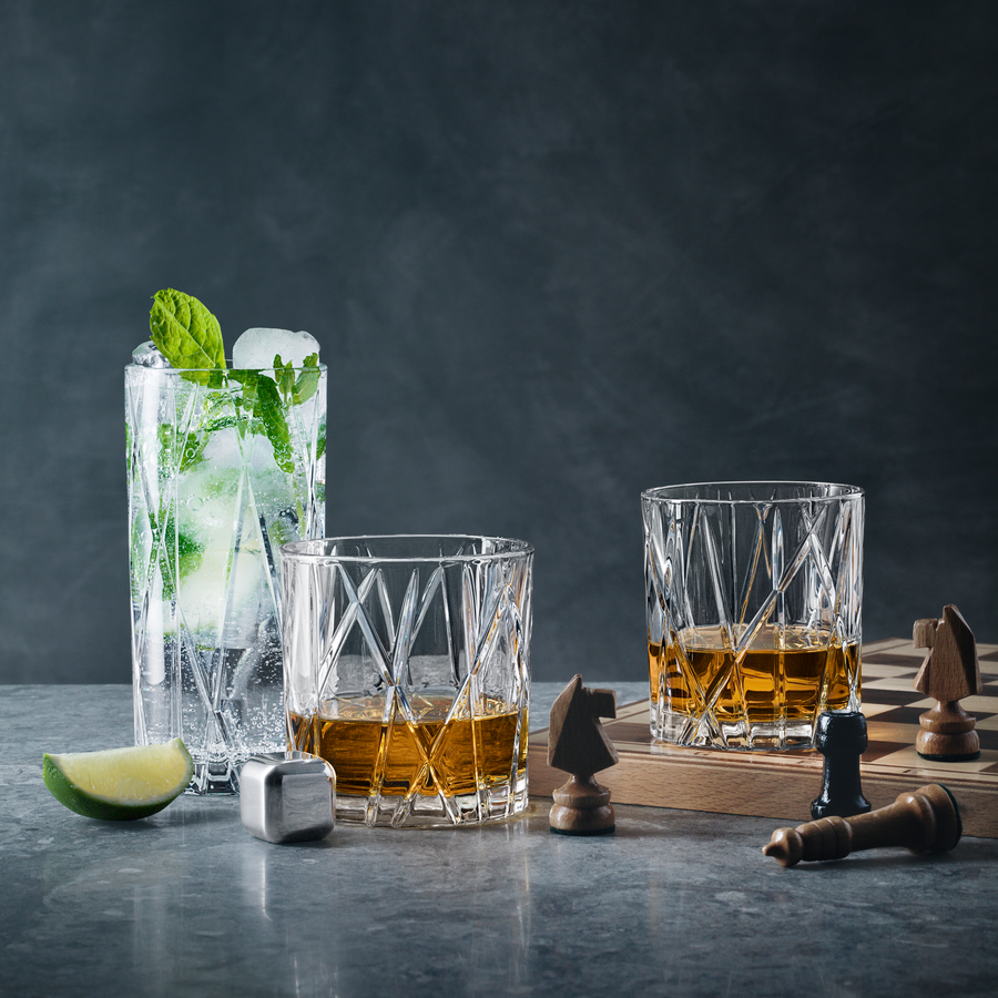 A tall mojito with mint and Orrefors: City Ice Cubes (4-Pack), two glasses of amber liquid, stainless steel-accented chess pieces, and a wooden board rest on a gray surface.