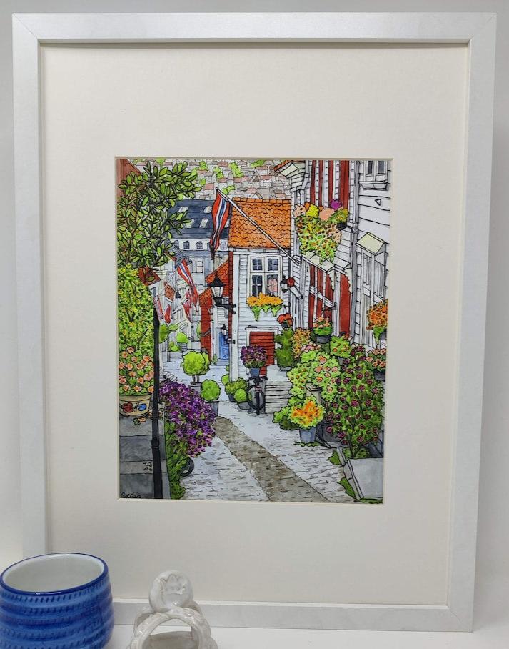 The product "Bergen Streets" by Greta Lann is a framed artwork depicting a charming Bergen street scene with colorful houses, blooming plants, and flags. A person strolling down the cobblestone road is shown alongside a blue cup and small sculpture, encapsulating the essence of Bergen, Norway.