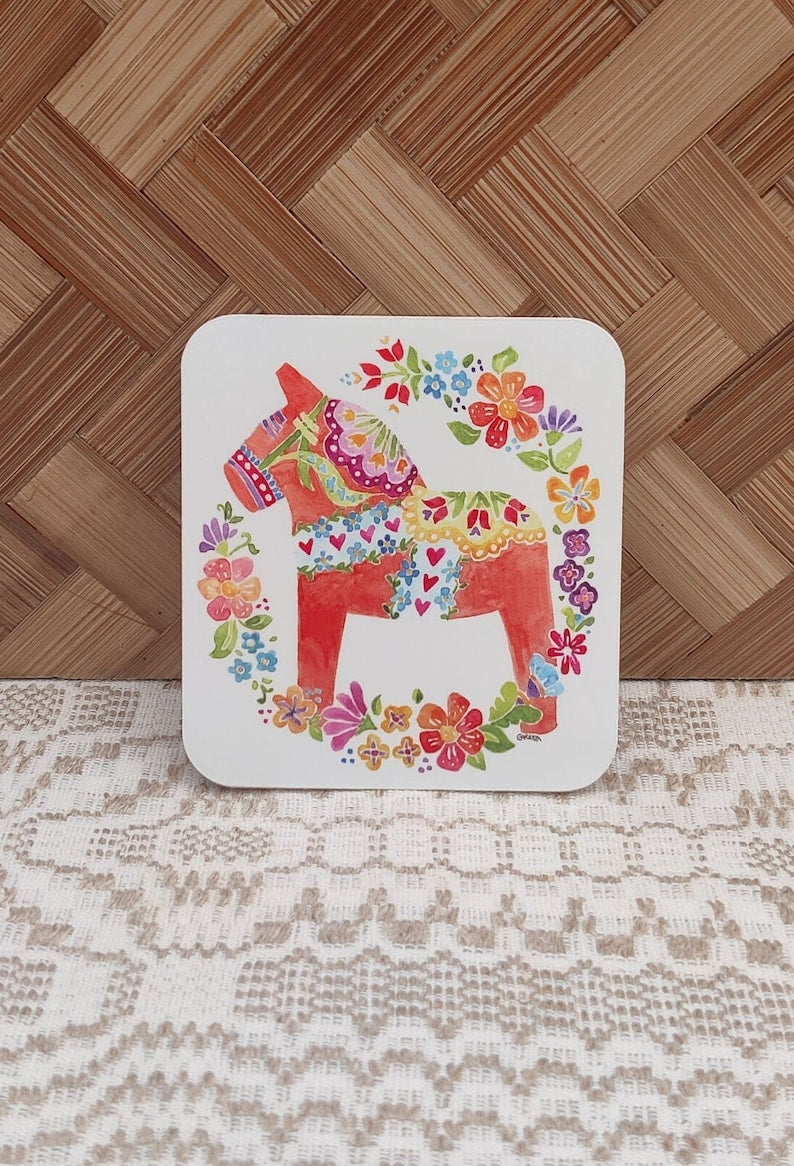 Sticker featuring a vibrant floral-patterned Dala Horse design by Greta Lann Custom, reminiscent of traditional Swedish art, set against a woven wooden background.