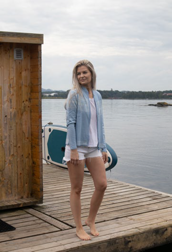 A woman in the Northern Story Setesdal Light Blue Norwegian Design Wool Cardigan stands barefoot on a wooden dock beside a small structure, with a paddleboard and calm water in the background.