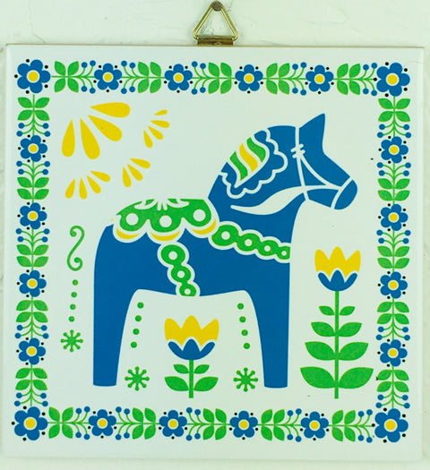 The 6" Ceramic Blue Dala Horse tile showcases a blue and green stylized Dala horse, adorned with yellow and blue flowers and encircled by a floral border.