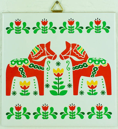 The 6" Tile: 2 Swedish Dala Horses and Swedish Flowers features two red Dala Horses facing each other, surrounded by green leaves and red and yellow floral patterns. It includes cork backing and a hanger for easy display.