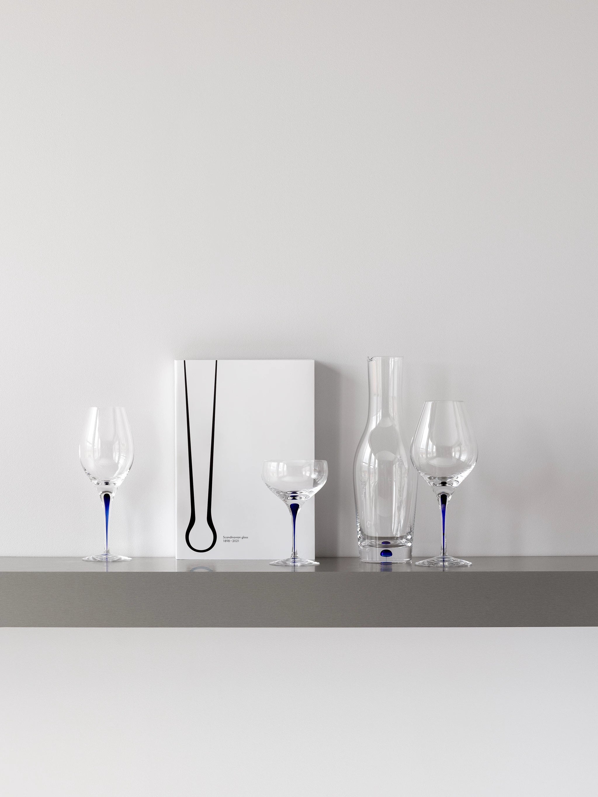 The minimalist shelf displays a book, an Orrefors glass vase, and three Orrefors Intermezzo Blue Wine Balance Glasses with striking blue stems against a pristine white wall.