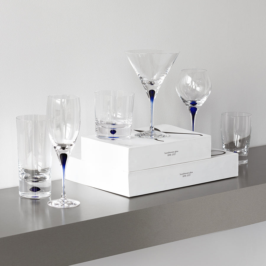 The Orrefors: Intermezzo Blue Martini Glass 8.5oz (2-Pack) by Erika Lagerbielke is elegantly displayed with clear and blue stemmed wine glasses and tumblers on white boxes and a gray shelf.