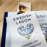 Three books rest on a wooden surface, with “Swedish Lagom: Finding Joy in Enough” atop two others. A Carl Larsson “Karin by the Shore” (8.5" × 2.5") bookmark peeks out, echoing classic Scandinavian artistry.