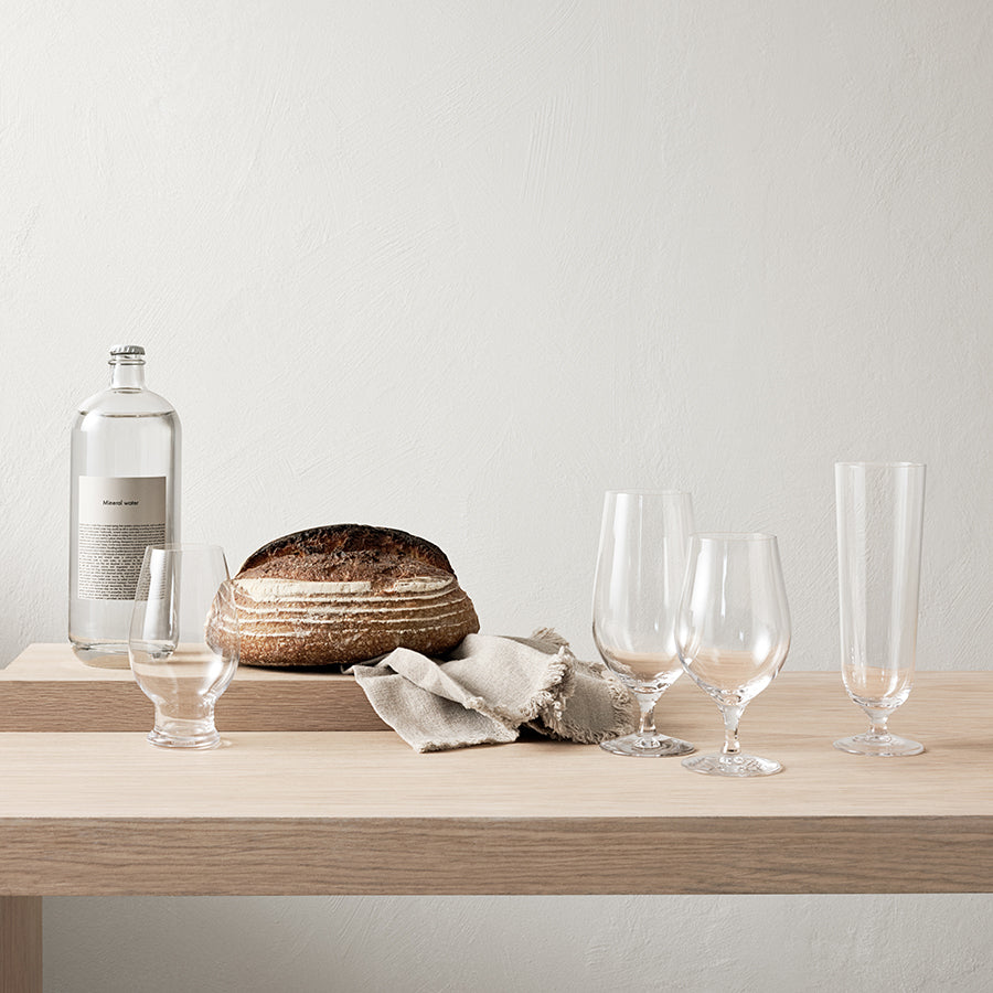 A loaf of bread, a bottle of clear liquid, a beige napkin, and four Orrefors Beer Lager 20oz glasses designed by Erika Lagerbielke are arranged on a light wooden table against a plain light-colored wall.