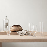 A loaf of bread, a bottle of clear liquid, a beige napkin, and four Orrefors Beer Lager 20oz glasses designed by Erika Lagerbielke are arranged on a light wooden table against a plain light-colored wall.