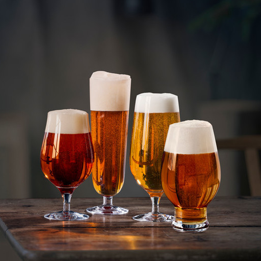 Four Orrefors Beer Lager 20oz glasses, designed by Erika Lagerbielke, sit on a wooden table. Each glass in the 4-pack features a tulip shape and holds a different shade of amber lager with a thick foam head.