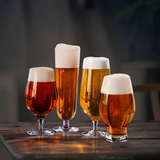 Four Orrefors Beer Lager 20oz glasses, designed by Erika Lagerbielke, sit on a wooden table. Each glass in the 4-pack features a tulip shape and holds a different shade of amber lager with a thick foam head.