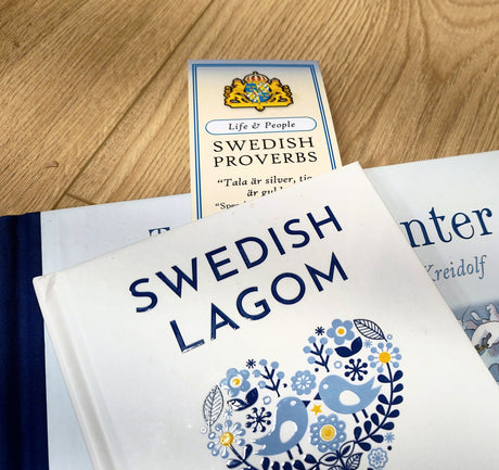 A book titled SWEDISH LAGOM rests atop another on a wooden surface, with the Bookmark: Swedish Proverbs “Life & People” (2.5" × 8.5") partially visible—offering a glimpse into Scandinavian wisdom.