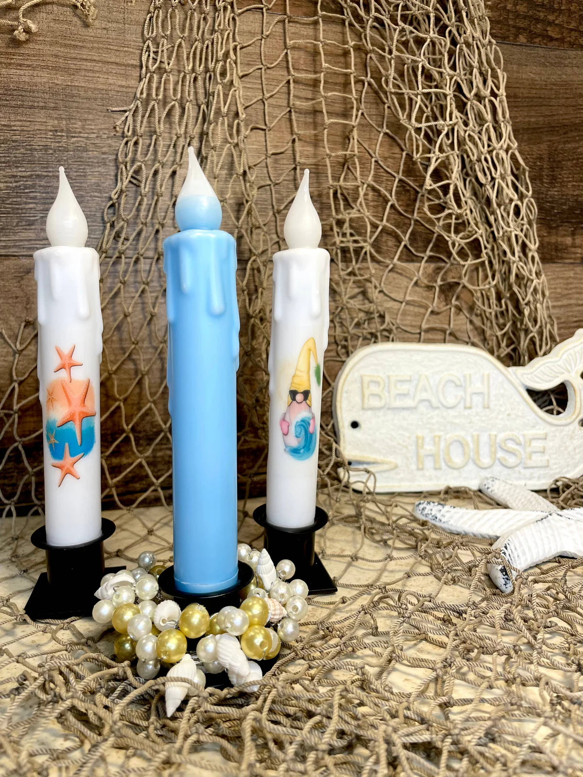 Three flameless candles, featuring a 7 tapered LED beeswax candle and two ocean-themed designs, are placed amid netting, pearls, shells, with a Beach House sign in the background.