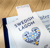 A book titled Swedish Lagom with a decorative cover rests atop another, evoking a calm Swedish summer. The Carl Larsson “Lisbeth Fishing” Bookmark (8.5" × 2.5") is partially inserted in the Swedish Lagom book.