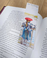 A book rests open on wood, marked by the Carl Larsson “Lisbeth Fishing” bookmark (8.5" × 2.5"), which showcases Lisbeth in her red hat and captures the essence of a Swedish summer.