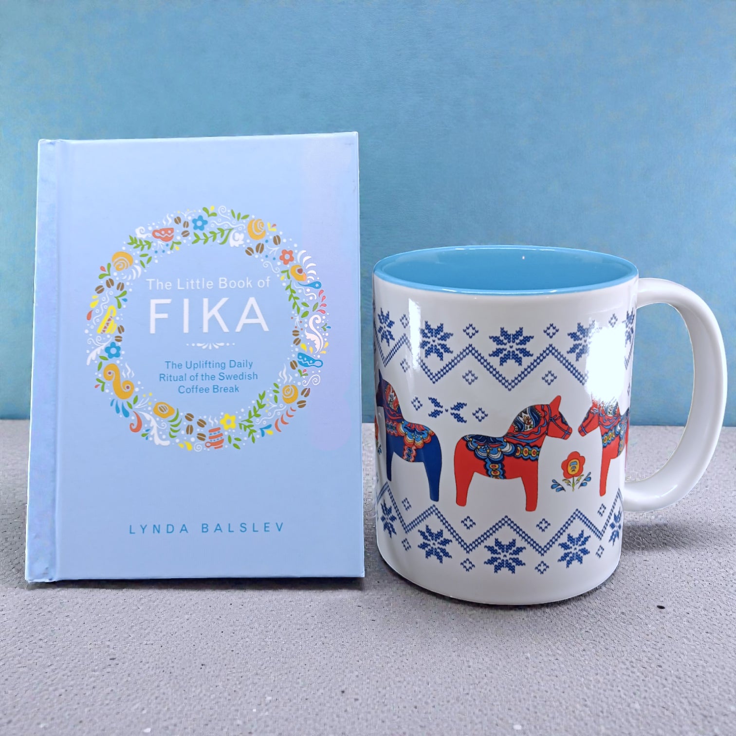 A light blue copy of "Little Book of Fika" by Lynda Balslev stands beside a white mug with blue patterns and red Dala horses, capturing the essence of a Swedish coffee break, all set against a blue background.