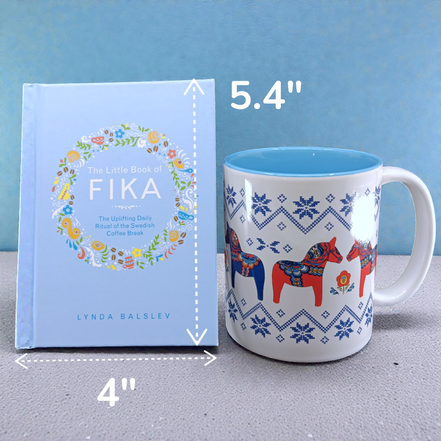 A light blue "Little Book of Fika" by Lynda Balslev stands beside a white mug with blue and red designs—the book measures 4 x 5.4", and the taller mug create an ideal Swedish coffee break setup with Scandinavian treats.