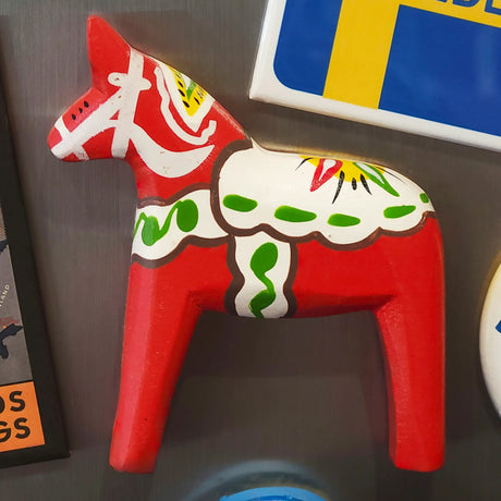 A red wooden Dala Horse magnet with painted white, green, and yellow decorative patterns, shown on a gray surface.