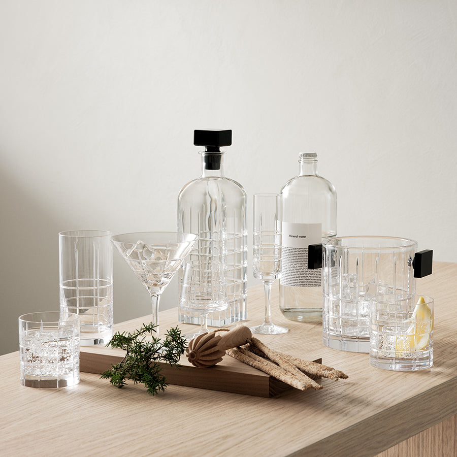 An exquisite collection of Orrefors glassware with a wooden tray of herbs and breadsticks rests on a light wood surface. A stylish Street Martini Glass by Orrefors, designed by Jan Johansson, stands beside tall and short glasses filled with ice.