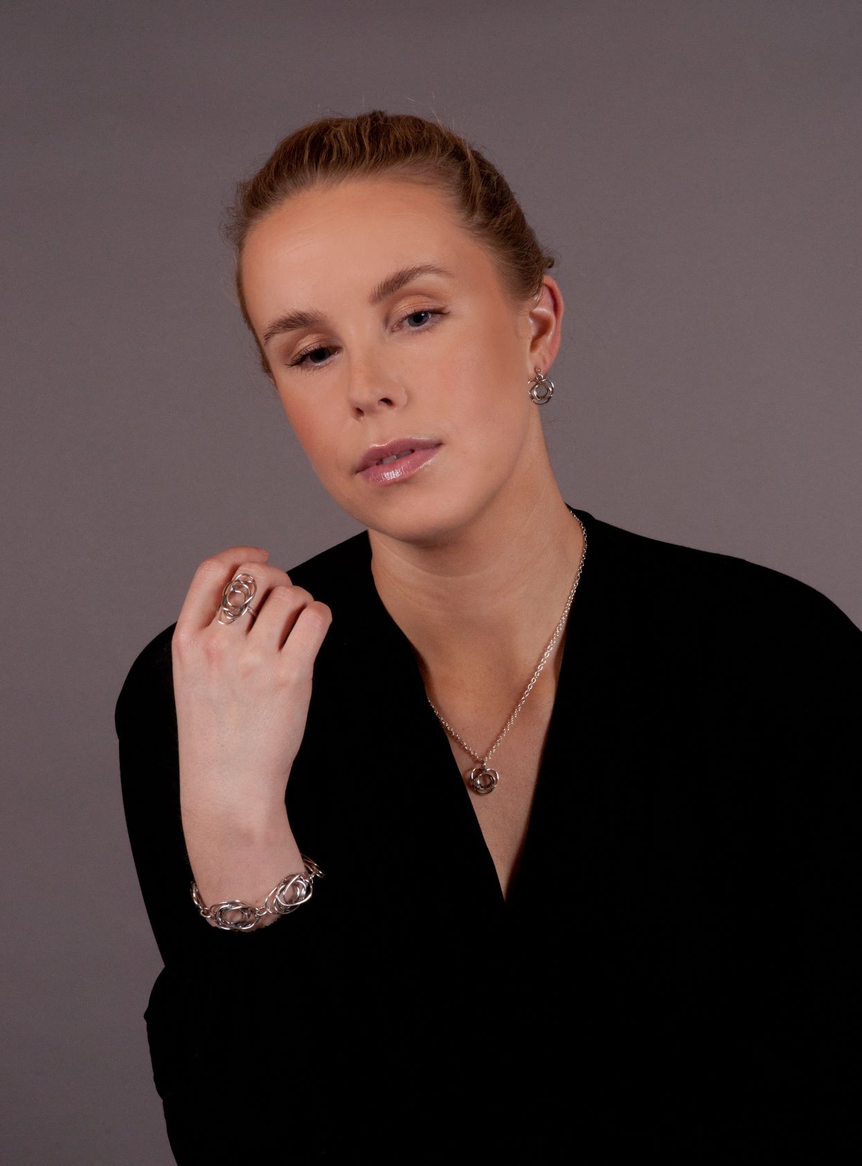 A woman with light skin and pulled-back hair wears a black top and the "Flor Alida" floral silver necklace by Hillestad (Norway), posing against a plain gray background.