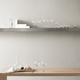 A minimalist dining scene with two Orrefors wine glasses, a decanter, and snacks on a wooden table; an array of glassware and Orrefors: More Champagne (4-Pack) 6 oz glasses line a floating shelf against a neutral wall.